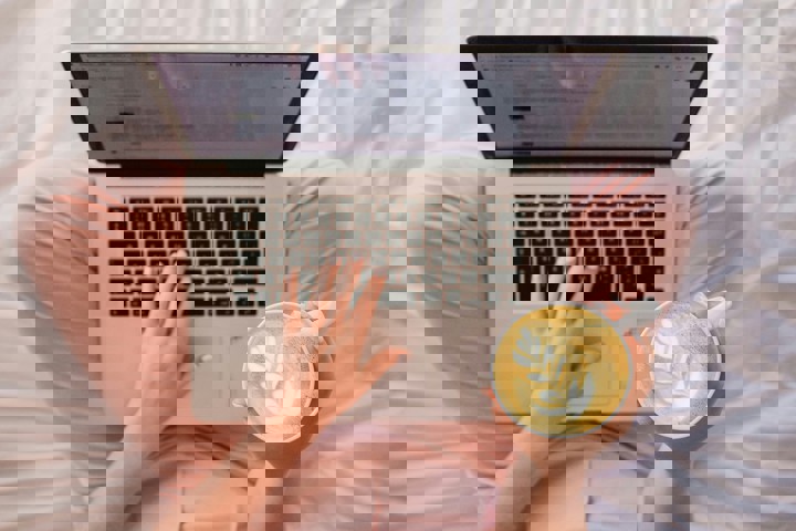 Laptop och kaffekopp i knäet på person med rosa byxor, sedd uppifrån